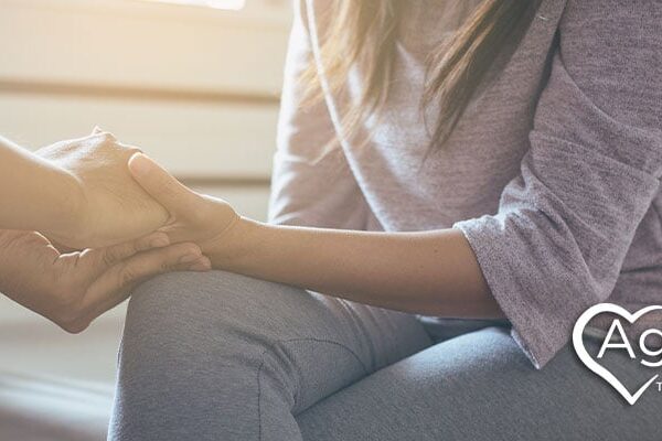 Person in gray sweater being comforted by another's hand. Agape Treatment Center logo with heart and dove outline.