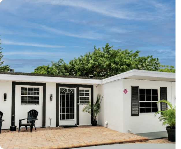 White house with black shutters, front porch seating, potted plants, and a sunny sky.