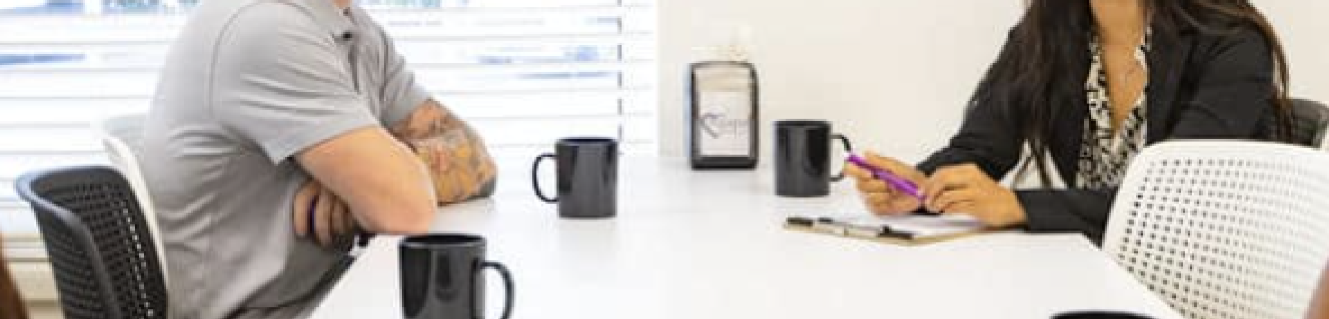 Two people sitting at a table, one in gray shirt with tattoos, the other in a black blazer and patterned shirt.