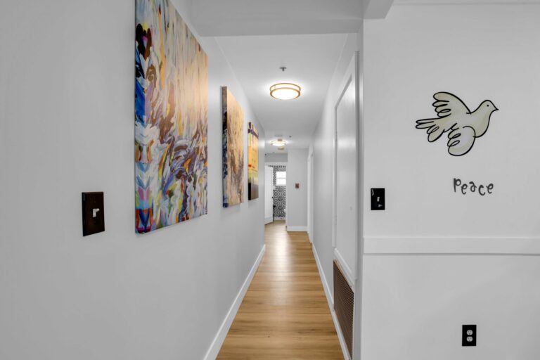Hallway with wooden floor, abstract paintings on left wall, dove drawing and Peace text on right wall.